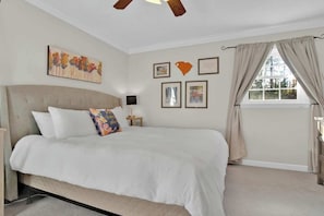 A bedroom with a large bed, a ceiling fan, a window with curtains, and several framed pictures on the wall.