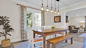 A dining area with a wooden table, white chairs, a bench, hanging pendant lights, a potted plant, and a window with patterned curtains.