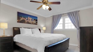 A hotel room with a bed, two bedside lamps, a ceiling fan, a window with curtains, and a painting on the wall.