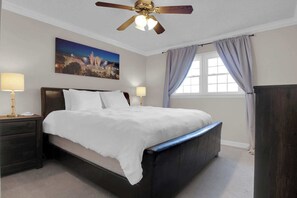 Quarto de hotel com cama, duas luminárias de cabeceira, ventoinha no teto, janela com cortinas e um quadro na parede.