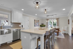 A modern kitchen with white cabinetry, stainless steel appliances, a central island with bar stools, pendant lighting, and a view of the outdoors through large windows.