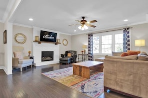 A living room with a fireplace, a ceiling fan, a television, and a large window with curtains.