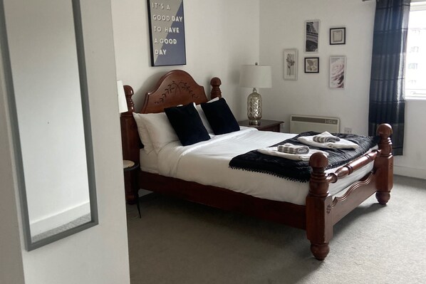 Una cama bien hecha con cabecera de madera, una mesita de noche con lámpara, y una pared decorada con cuadros y una cita motivacional.