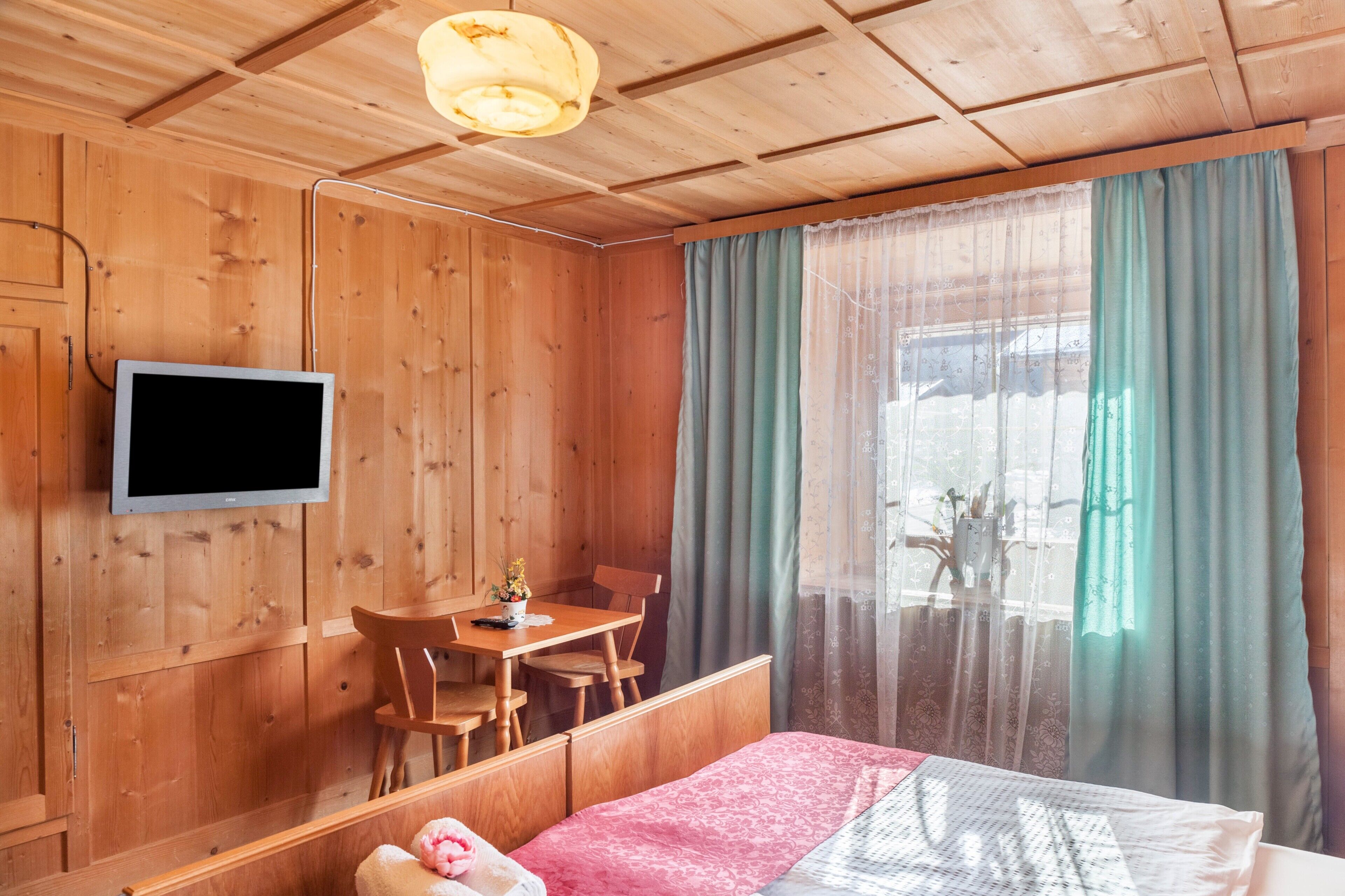 Ein Zimmer mit Holzboden, einem Bett, einem Fernseher, einem Esstisch mit Stühlen und einem Fenster mit Vorhängen.