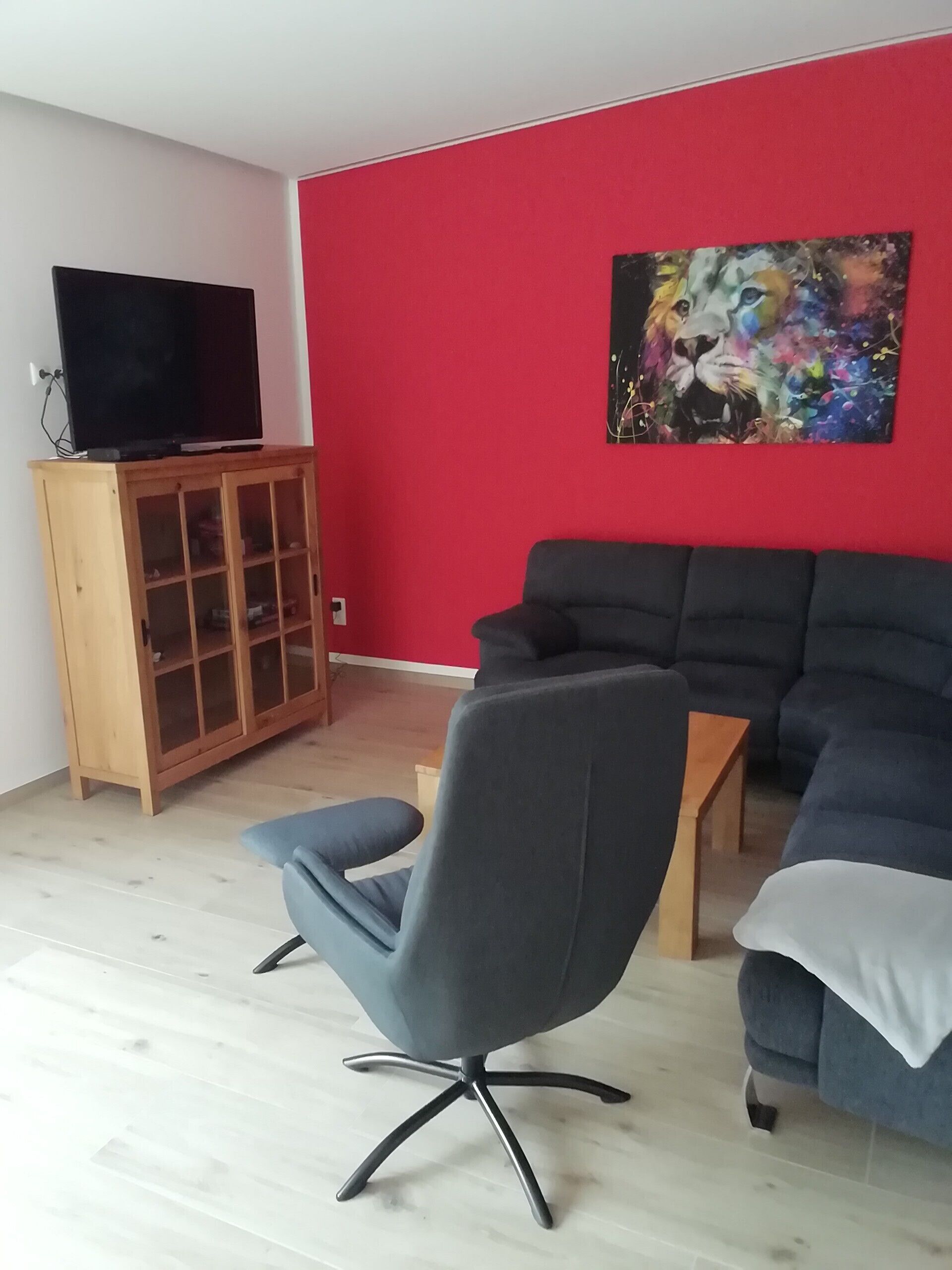 Un salon avec un mur rouge, un canapé noir, un fauteuil de bureau gris, un meuble en bois sur lequel est posée une télévision, et un tableau.