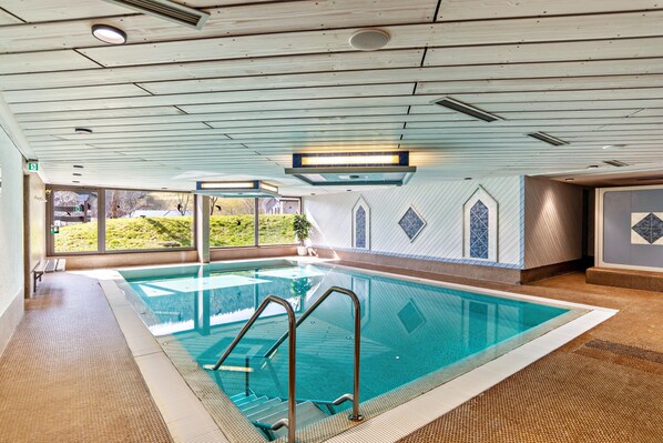 An indoor swimming pool with clear glass walls, a wooden ceiling, and a view of greenery outside.
