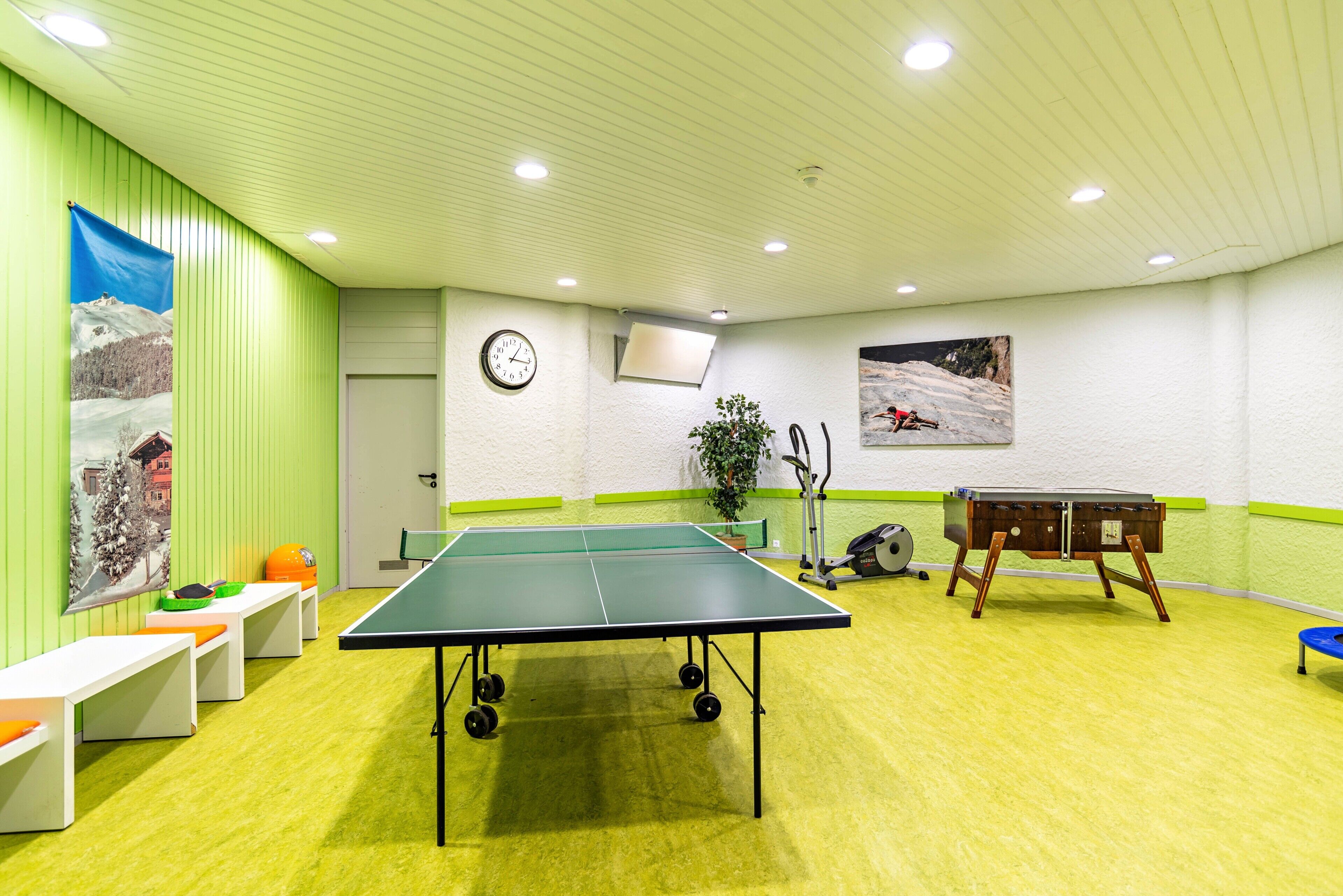 Une pièce avec une table de ping-pong, des bancs, une horloge, une plante et une table sur laquelle se trouve un sac.
