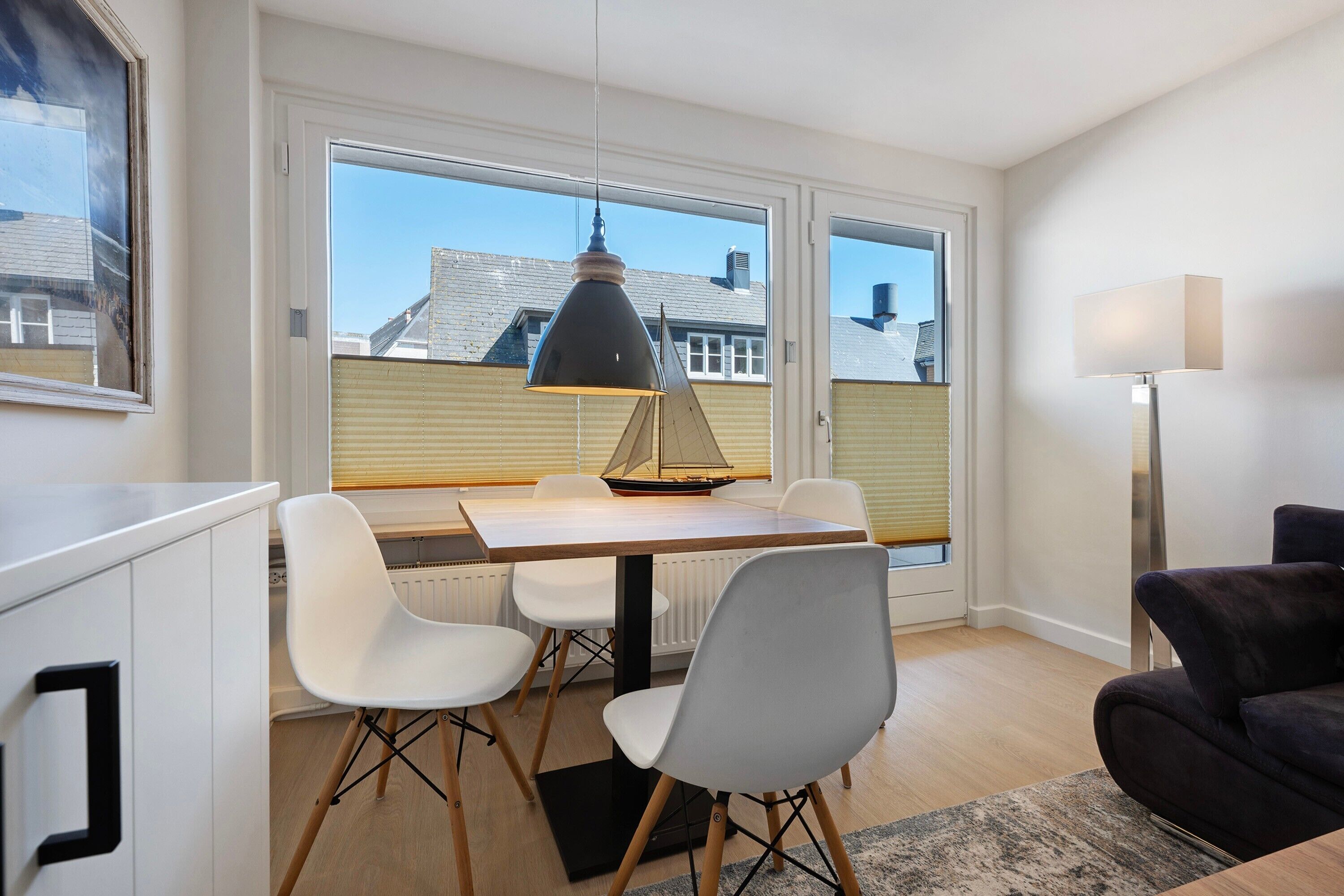 A dining area with a table, chairs, a pendant light, and a view through large windows.