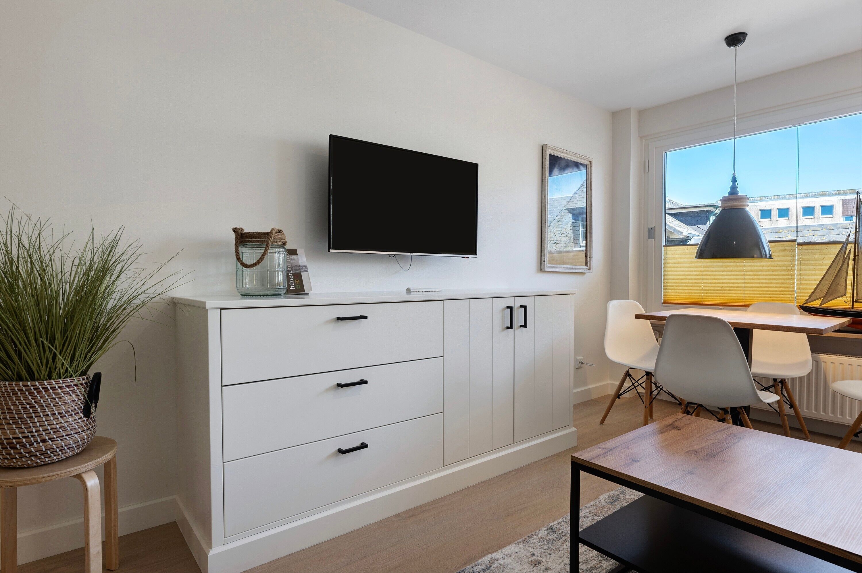 En moderne stue med et fladskærms-tv monteret på væggen, et spiseområde med bord og stole, og et stort vindue med udsigt til bygninger.