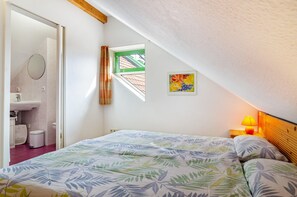 A bedroom with a bed, a window, a small wall-mounted picture, and a nightstand with a lamp.