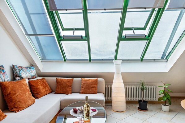 A modern living room with a glass ceiling, a sofa with brown cushions, a glass coffee table with a bottle and glasses, and two potted plants.