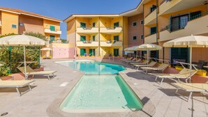 A pool area with lounge chairs and umbrellas, surrounded by a building with balconies and a clear blue sky.