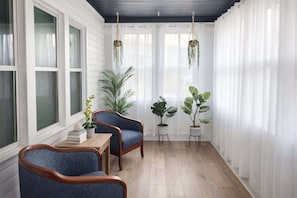 Un solárium con paredes blancas, piso de madera, dos sillones azules, una mesita con libros y plantas colgantes.