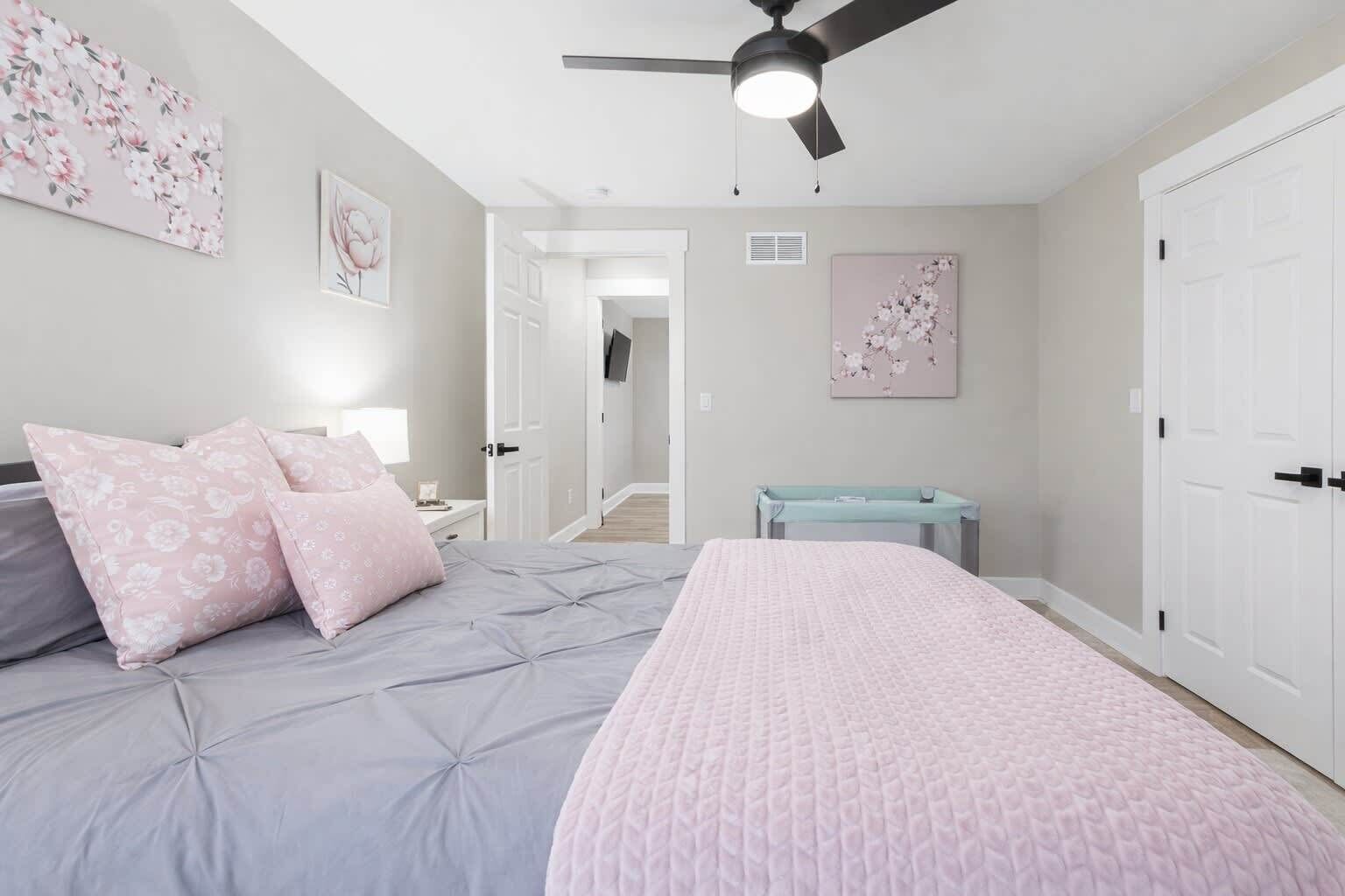 A bedroom with a bed, pillows, a ceiling fan, a nightstand, a desk, a chair, and a door.
