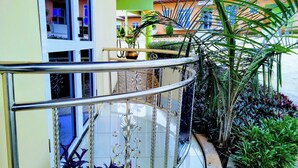 A curved glass balcony with metal railings, surrounded by potted plants and a view of a building with a sign.