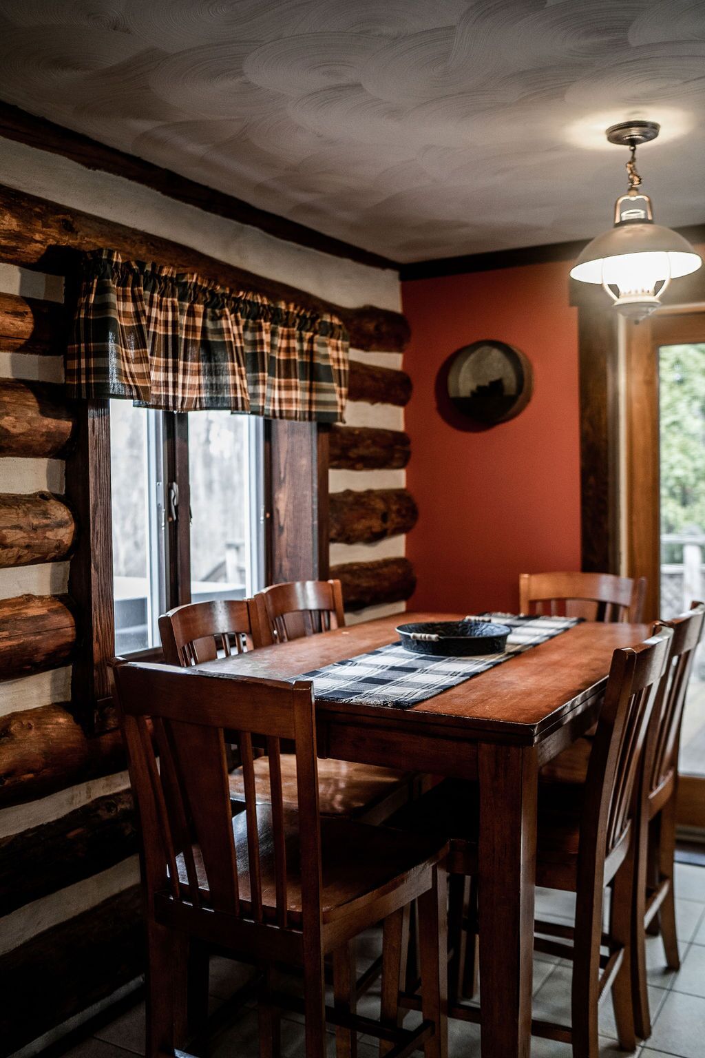 A rustic dining area with a wooden table and chairs, a plaid window valance, and a round mirror on the wall.