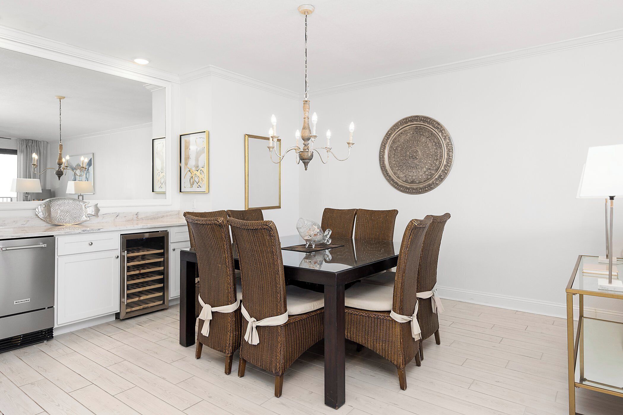 A dining area with a dark wooden table, four wicker chairs with white cushions, a chandelier, and a large mirror on the wall.