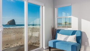 Una habitación con un amplio ventanal corredero que da a una vista de la playa, un sofá azul con un cojín blanco, y un ventanuilla con vista al mar.