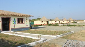 A row of thatched-roof huts with a central paved walkway, surrounded by a grassy area with a few plants. - Ram Rann Resort (Bhachau)