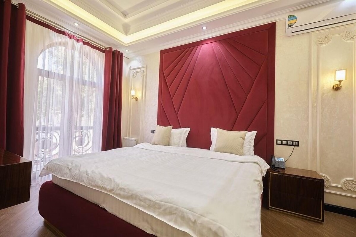 Une chambre d’hôtel avec un grand lit, une tête de lit rouge et une vue sur l’extérieur à travers des rideaux transparents.