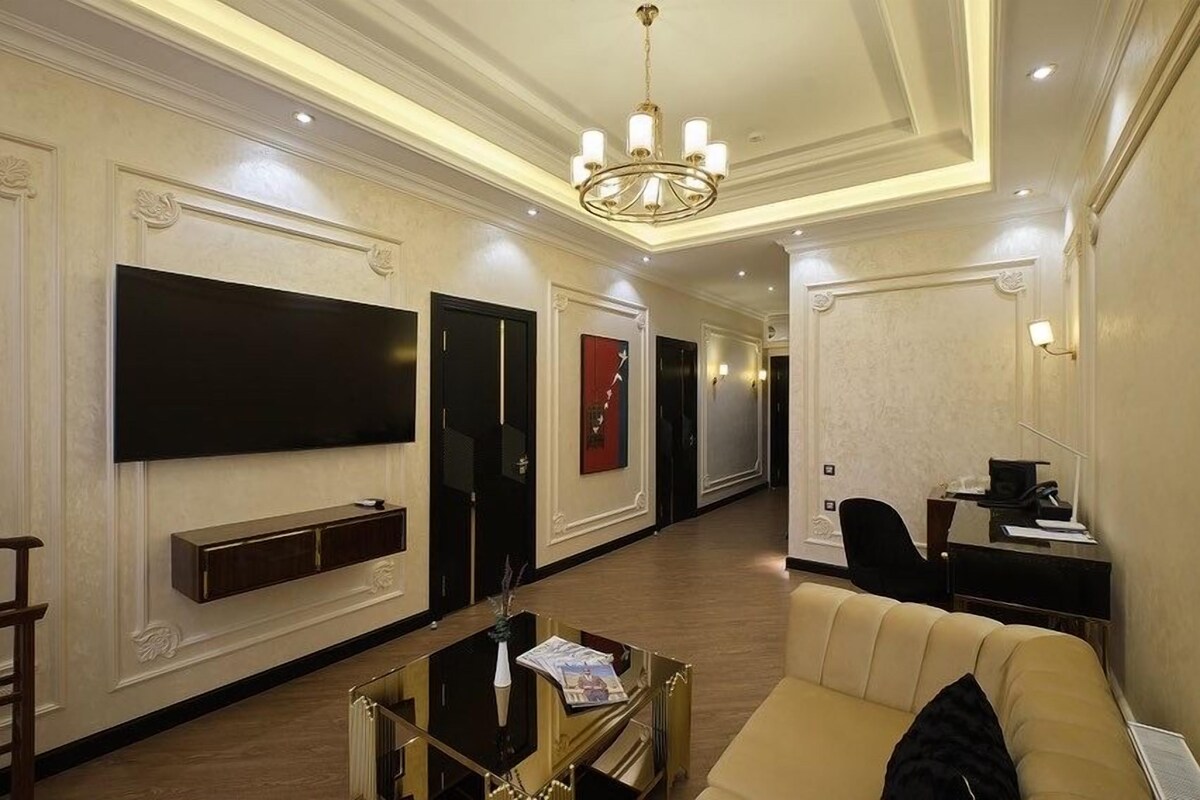 A hotel lobby with a sofa, a TV mounted on the wall, a chandelier, and a desk with a chair.