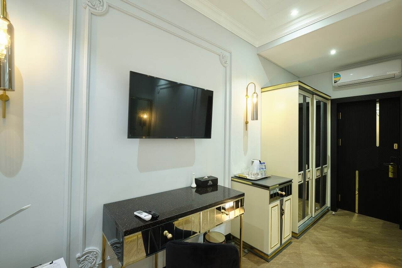 A hotel room with a flat-screen TV mounted on the wall, a small desk with a chair, a minibar, and a sliding door leading to another room.