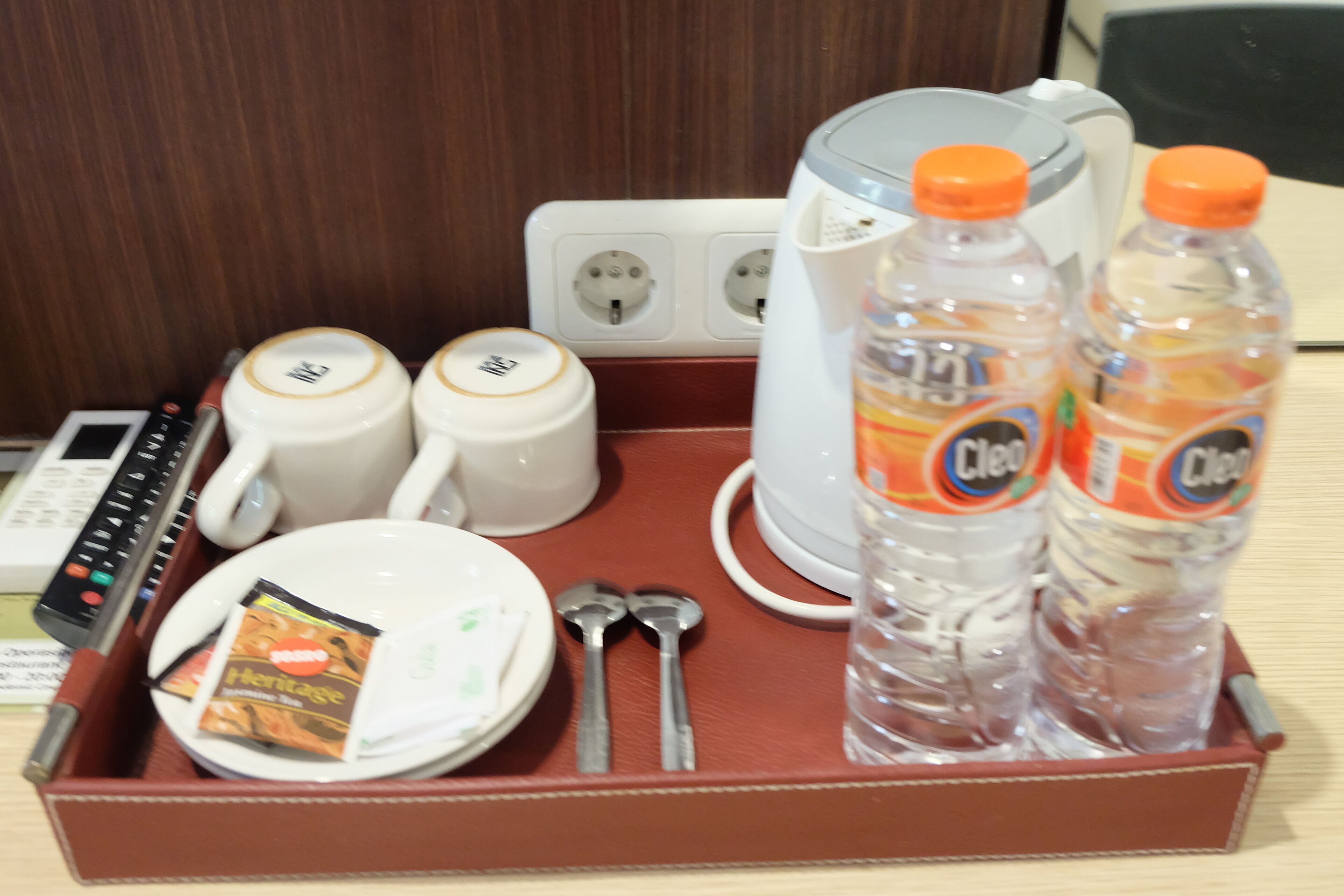 A hotel room's breakfast tray with a coffee maker, two mugs, a plate with a packet, two spoons, two bottles of water, and a remote control.