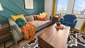 Uma sala de estar moderna com um sofá cinzento, almofadas coloridas, uma mesa de centro de madeira e uma poltrona azul. Um mapa está exposto na parede, e uma janela oferece vista para a cidade.