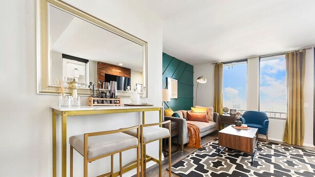 Quarto de hotel moderno com um espelho grande, um espaço bar com banquetas, um sofá, uma mesa de centro e vista do céu através de amplas janelas.