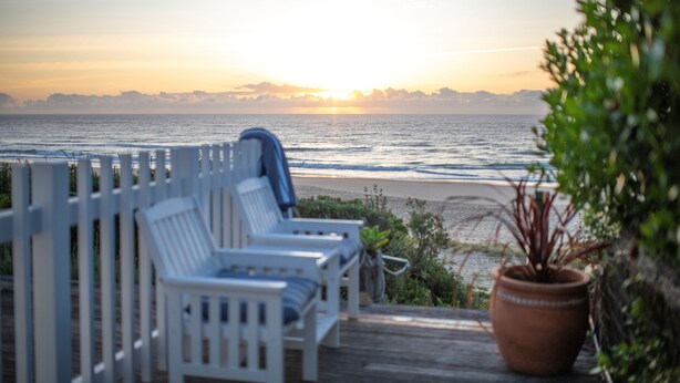 Eine Terrasse mit weißen Stühlen mit Blick auf einen Strand bei Sonnenuntergang.