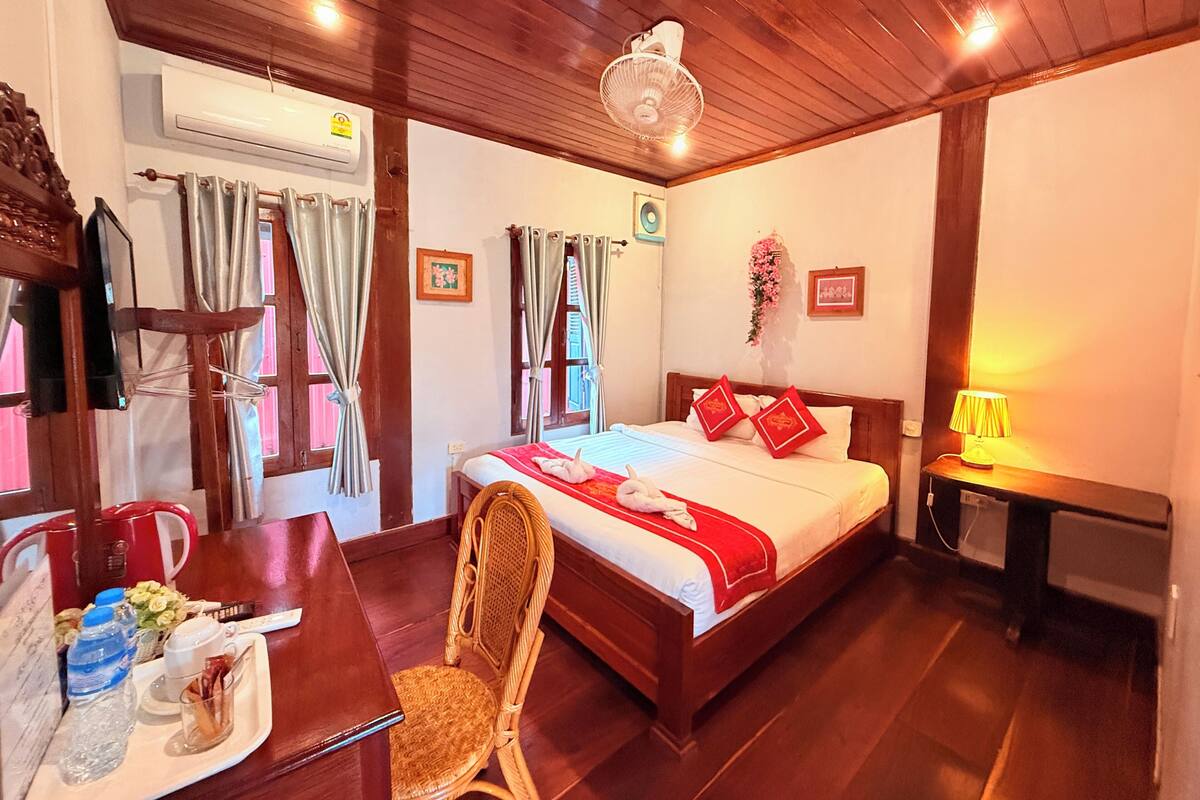 A hotel room with a wooden ceiling, a bed with red and white bedding, a bedside table with a lamp, a chair, a desk with a coffee maker, and a window with curtains.