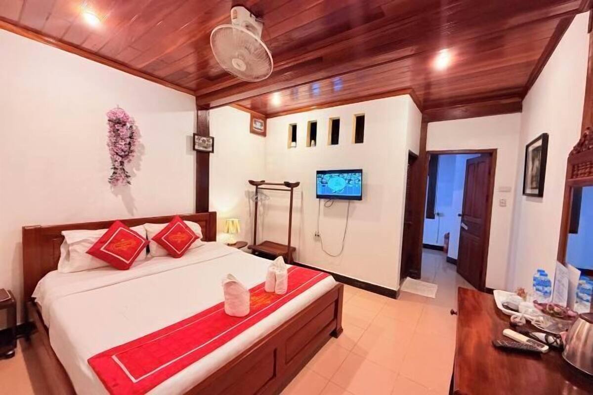 A hotel room with a wooden ceiling, a bed with red pillows, a TV mounted on the wall, and a shower area.