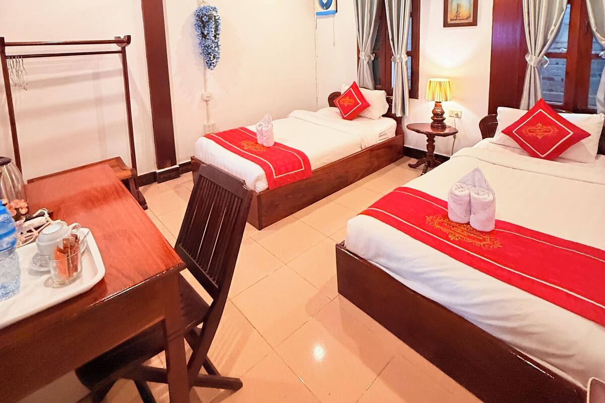 A hotel room with two beds, a desk, a chair, a small table with a lamp, and a window with curtains.