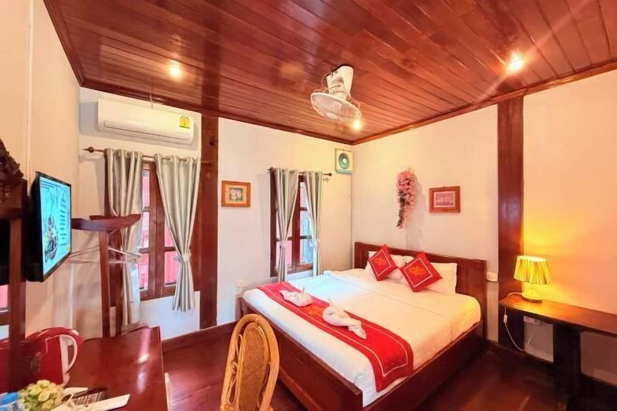 A hotel room with a wooden ceiling, a bed with red bedding, a TV, and a desk with a chair.