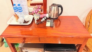 Une table en bois sur laquelle se trouve un plateau contenant une tasse de café, un verre d’eau, une bouteille d’eau, un petit panier de fleurs, une bouilloire, une télécommande et un adaptateur secteur.