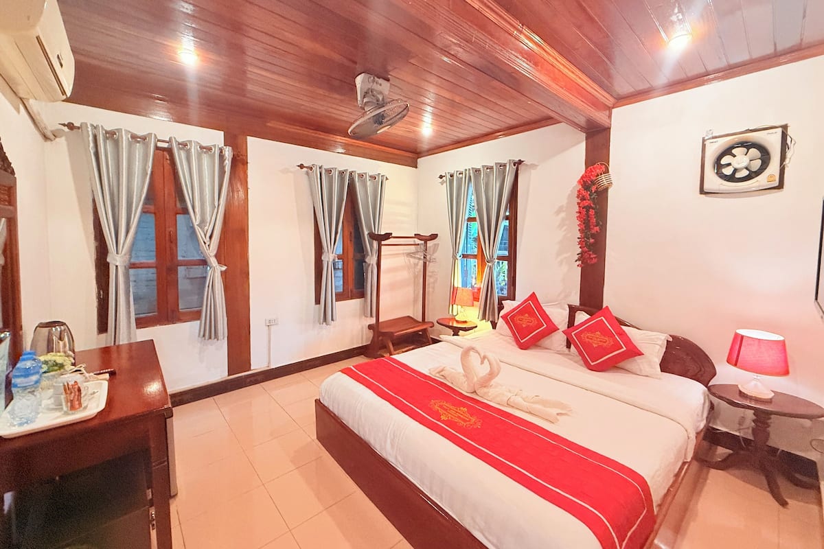 A hotel room with a bed, a desk, a chair, a small table, and a wall-mounted fan.
