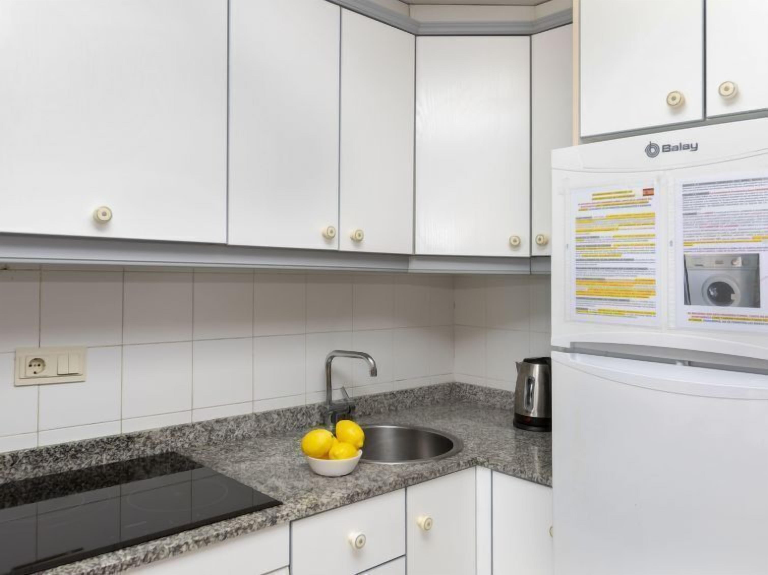 A kitchen with white cabinets, a granite countertop, a sink, a kettle, and a refrigerator with a manual.