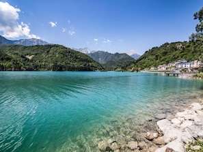 Un lago tranquilo de aguas claras con una orilla rocosa, rodeado de colinas y montañas cubiertas de vegetación exuberante.