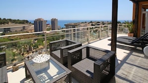 Un balcón con mobiliario de mimbre, una mesa de vidrio y vista al mar y edificios.