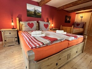 Una cama con ropa de cama a cuadros y almohadas con estampado de corazones, una mesita de noche de madera con una lámpara, y un cuadro en la pared.