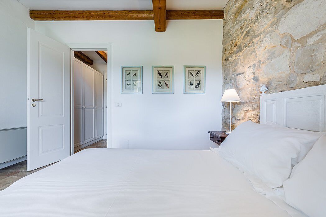 Un dormitorio con pared de piedra, cama con ropa de cama blanca, mesita de noche con lámpara y tres cuadros enmarcados en la pared.