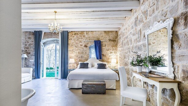 A bedroom with a stone wall, a large bed, a chandelier, and a view of the outdoors.