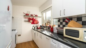 A compact kitchen with white cabinets, a black countertop, a microwave, a dishwasher, and a window with a view. - Super 1BD Garden Flat Islington London (London)