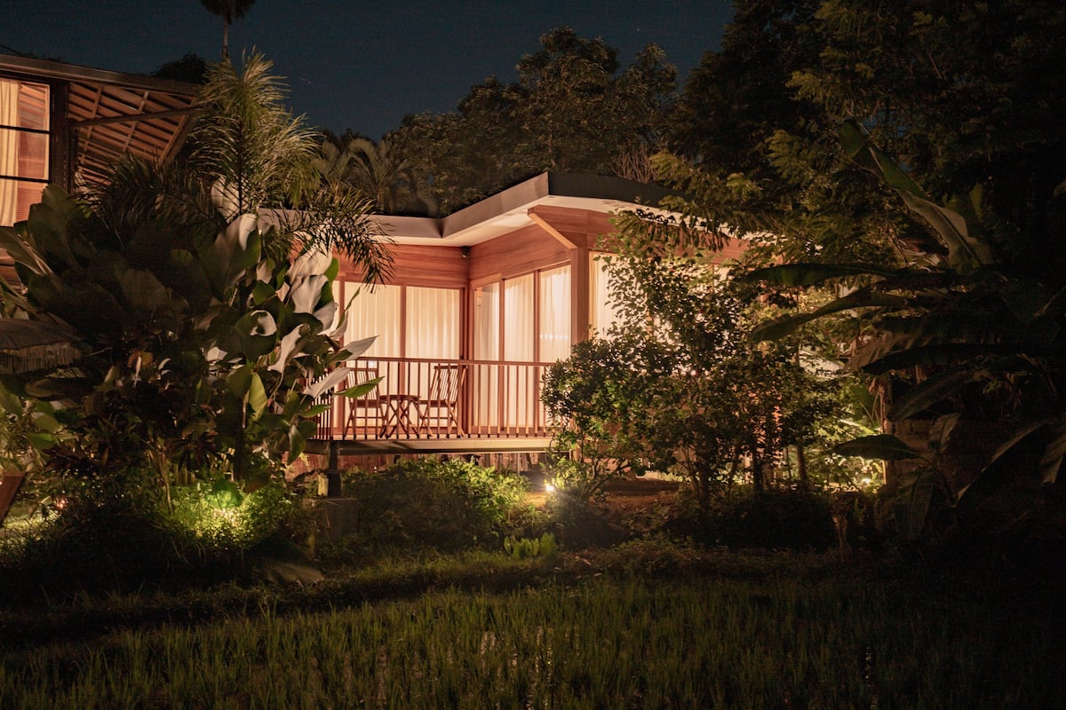 A house with a wooden deck, surrounded by lush greenery and palm trees.