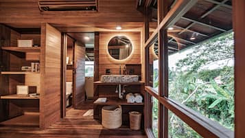 A wooden interior with a wooden floor, a circular mirror, a sink, and a window overlooking greenery.
