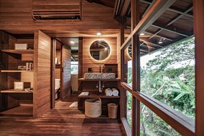 Un interior de madera con piso de madera, un espejo circular, un lavamanos y una ventana con vistas a follaje.