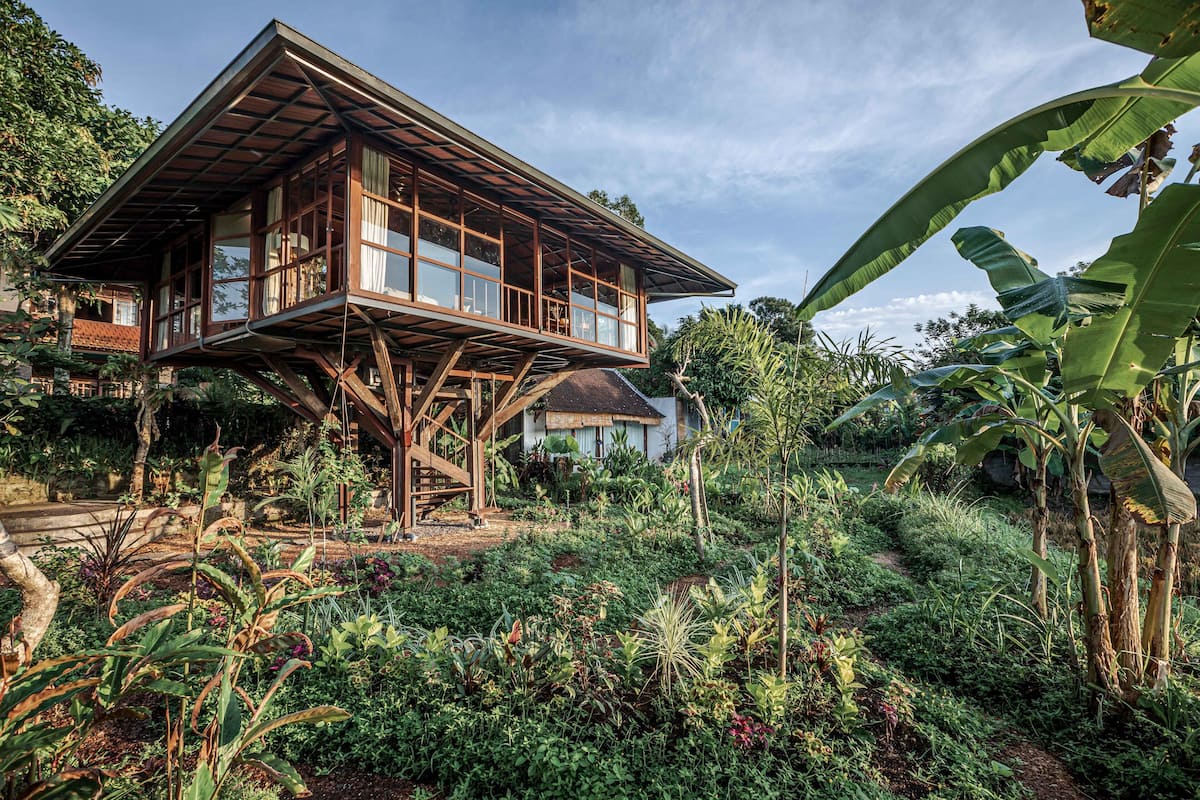A wooden house with a balcony, surrounded by lush vegetation and banana trees.