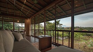 A spacious room with a large sofa, a wooden table, and large windows offering a view of a lush landscape. - Grün Ubud (Ubud)