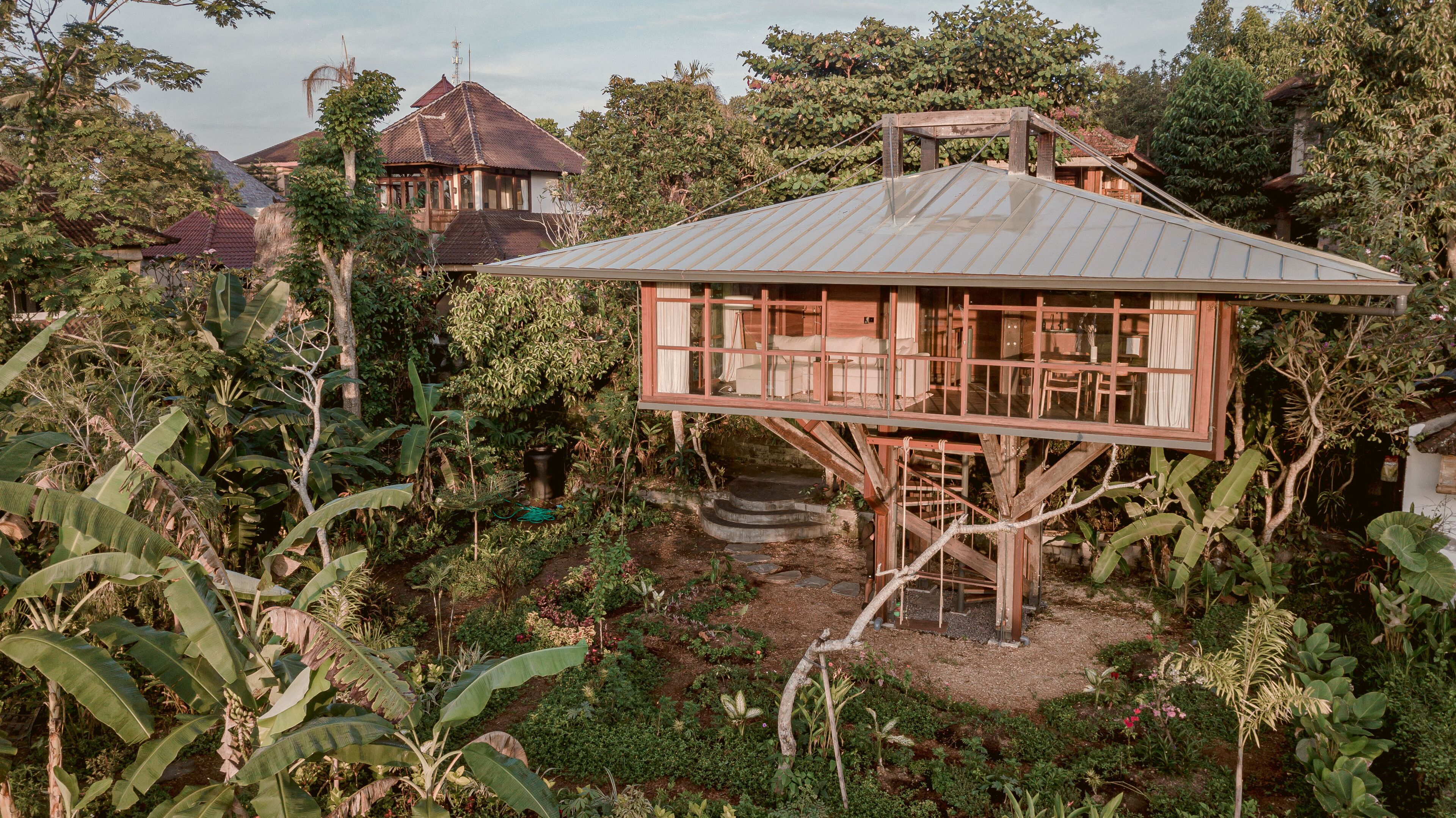 A treehouse with a wooden deck and a metal roof, surrounded by lush vegetation and a garden.