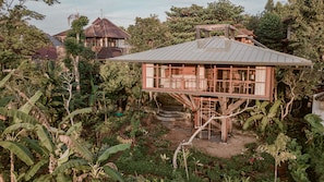 Una casa en los árboles con terraza de madera y techo metálico, rodeada de exuberante vegetación y un jardín.